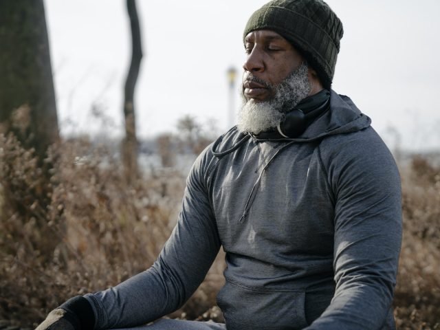 Bearded African American male in warm activewear and knitted hat sitting and meditating with closed eyes in autumn nature